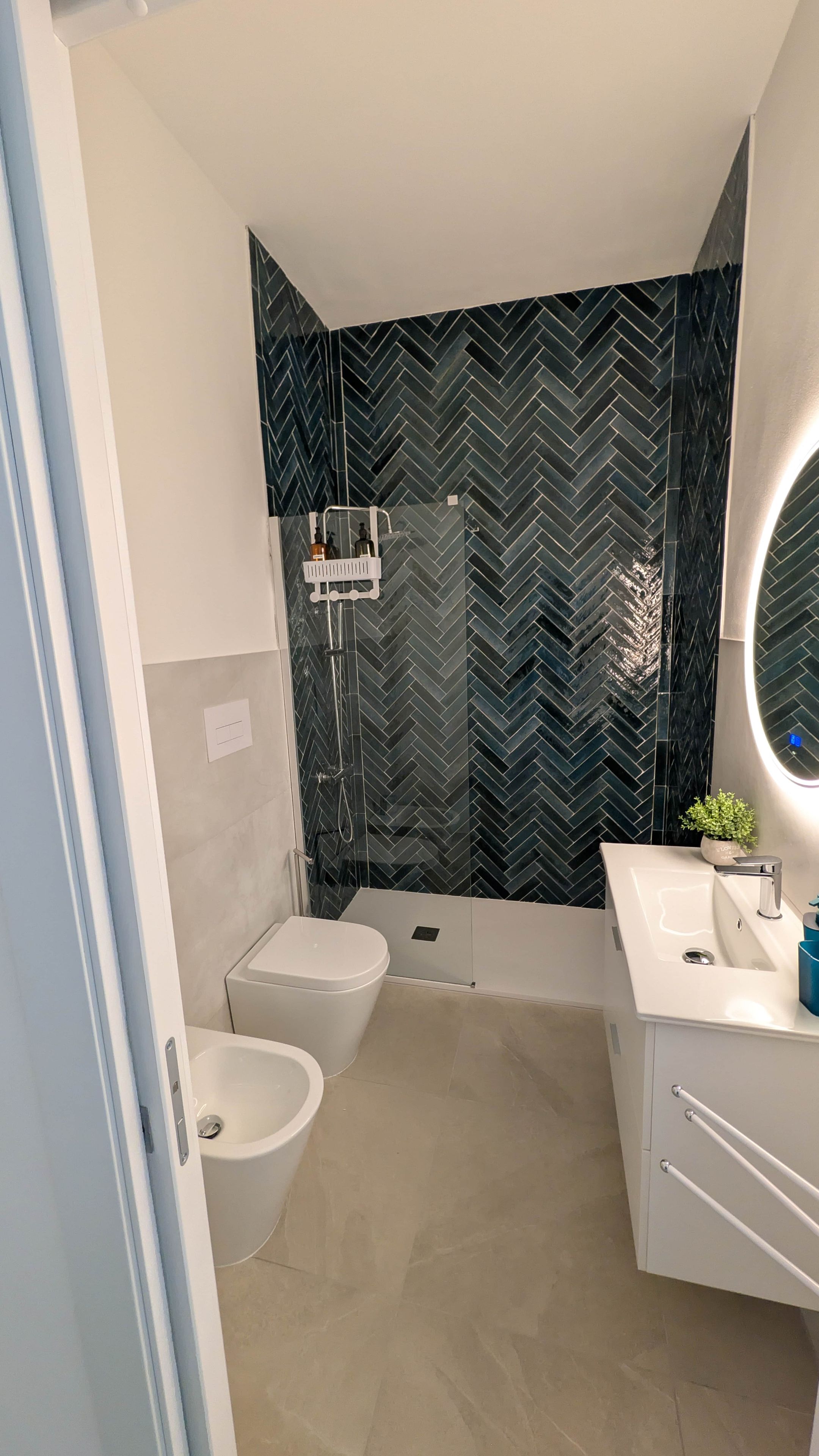 Une salle de bain moderne avec un mur recouvert de carreaux disposés en motif chevron, des toilettes blanches, un bidet, une baignoire et un meuble-lavabo avec un miroir.