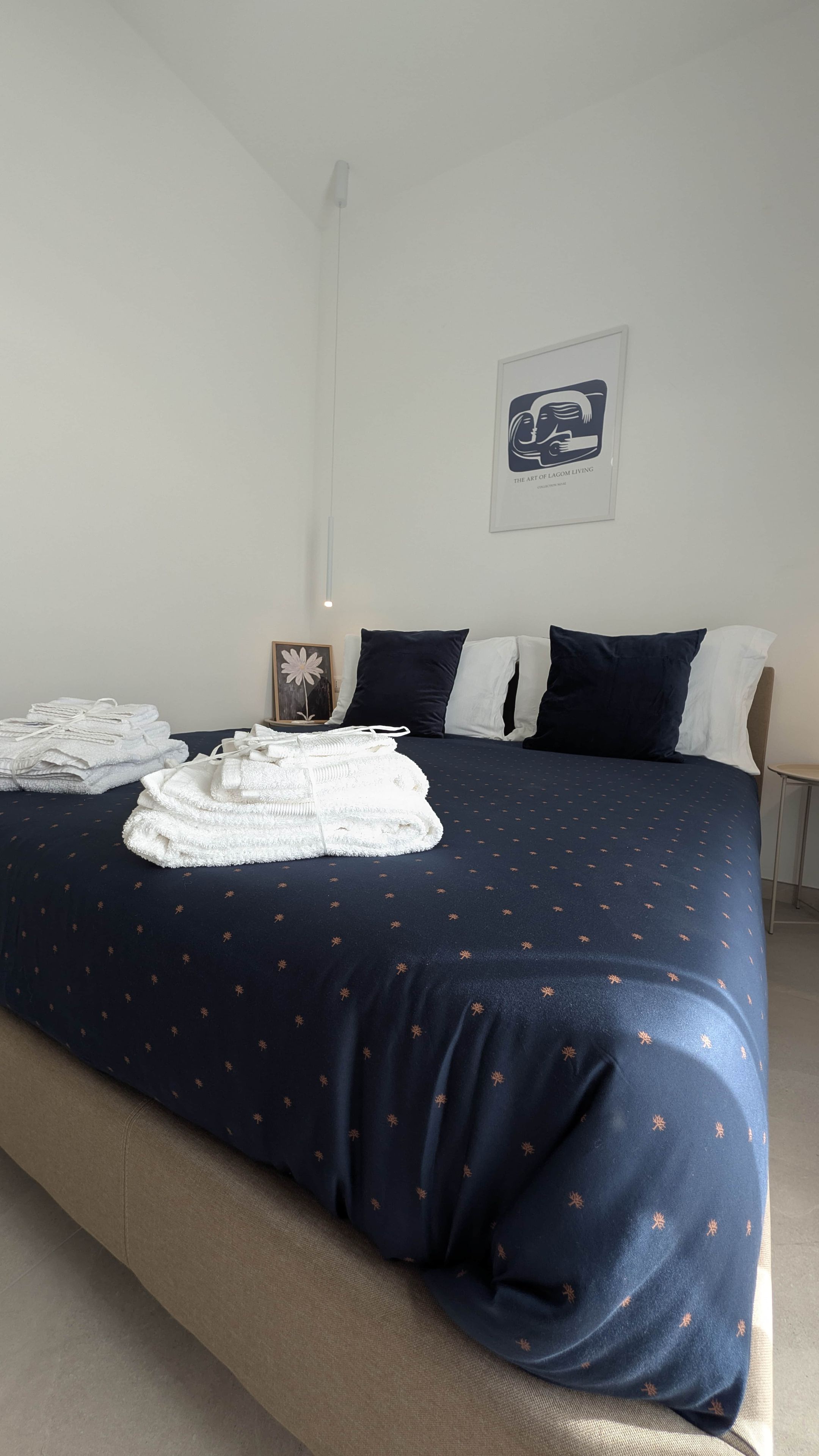 Un letto rifatto con una coperta blu scuro e asciugamani bianchi, un quadro appeso al muro e un comodino con una lampada.