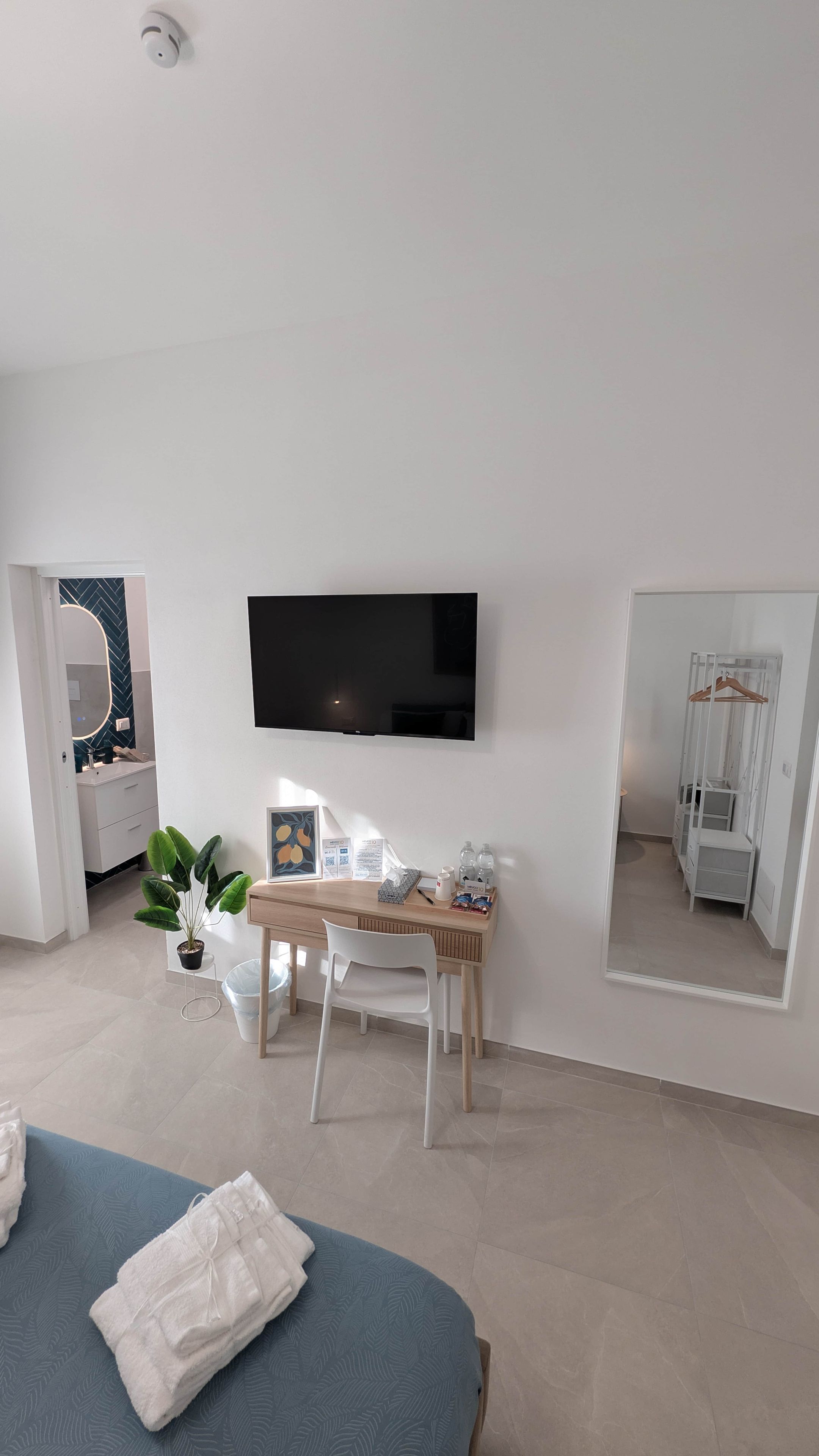 Una habitación de hotel moderna con cama, escritorio con silla, televisor de pantalla plana en la pared, espejo y armario con perchero.