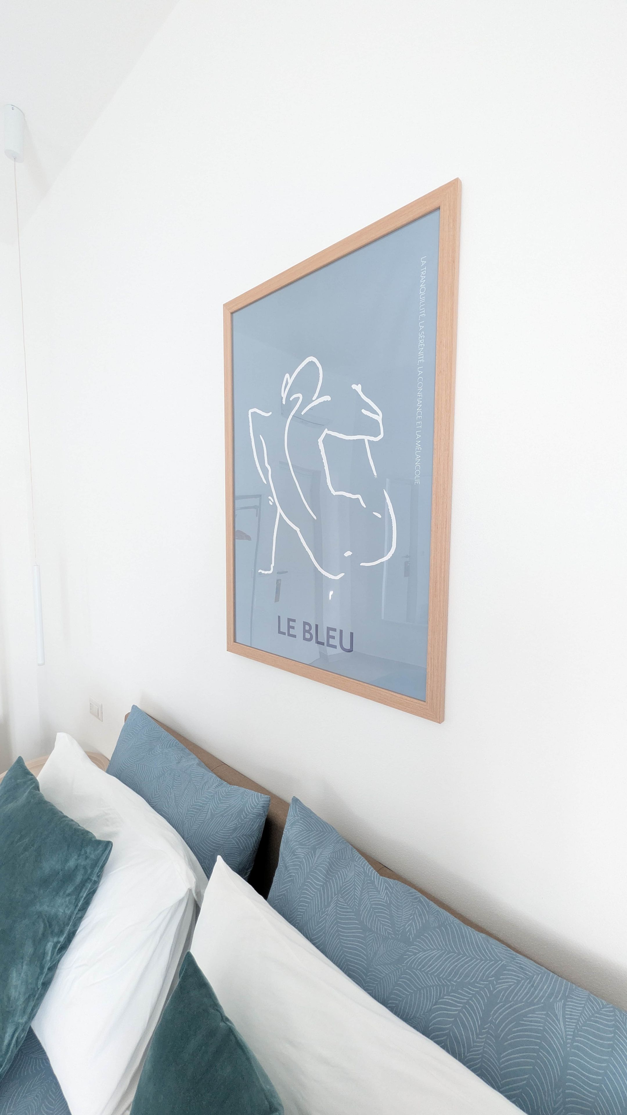 Ein gerahmtes Kunstwerk mit schlichtem Design, ein Bett mit gemusterten Kissen und eine weiße Wand.