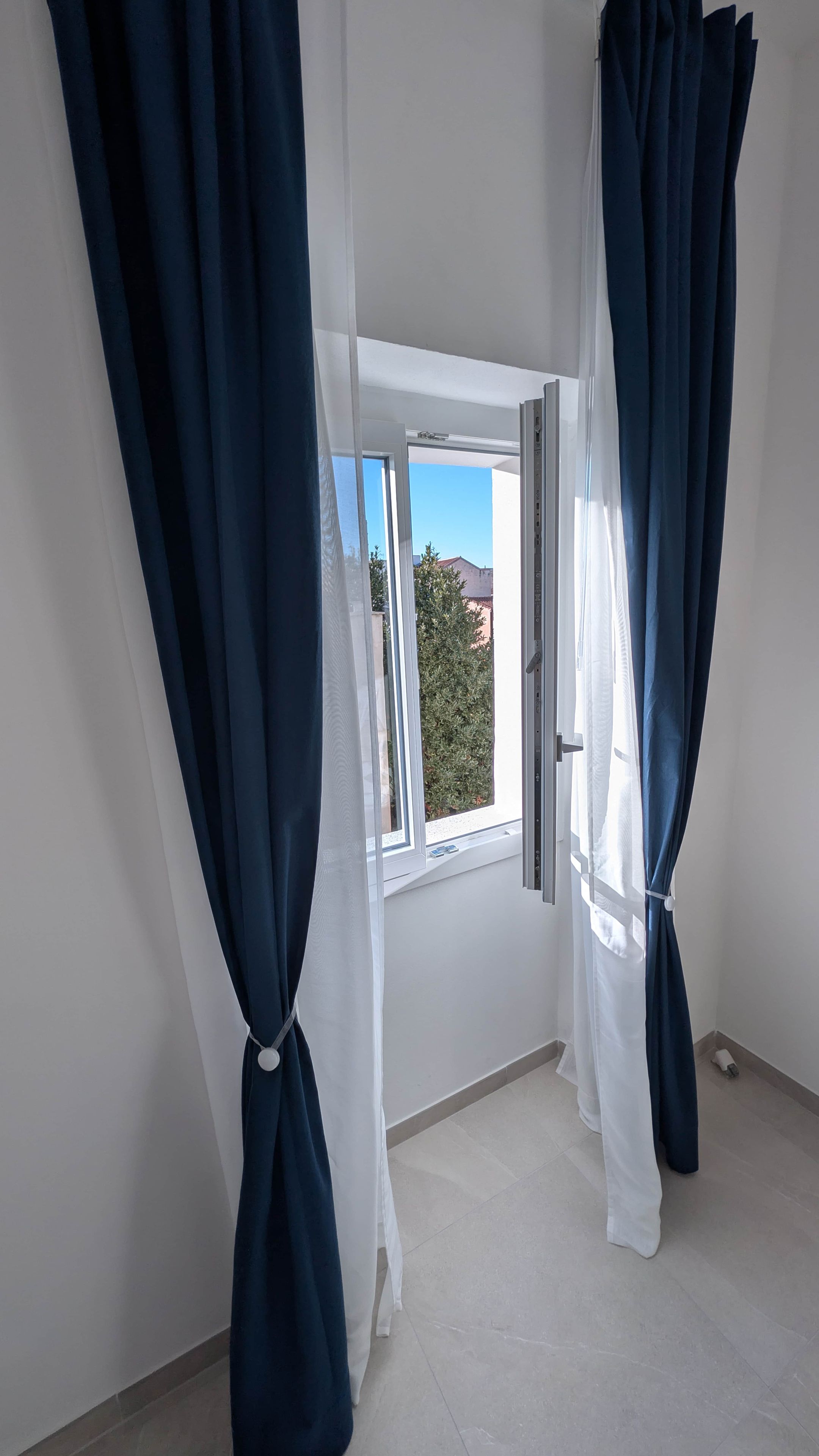 A room with a large window, white curtains, and a view of greenery outside.