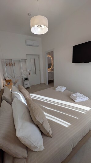 Ein modernes Schlafzimmer mit Bett, Kissen, einem Nachttisch, einem Fernseher und einem durchgehenden Türrahmen einsehbaren Badezimmer.