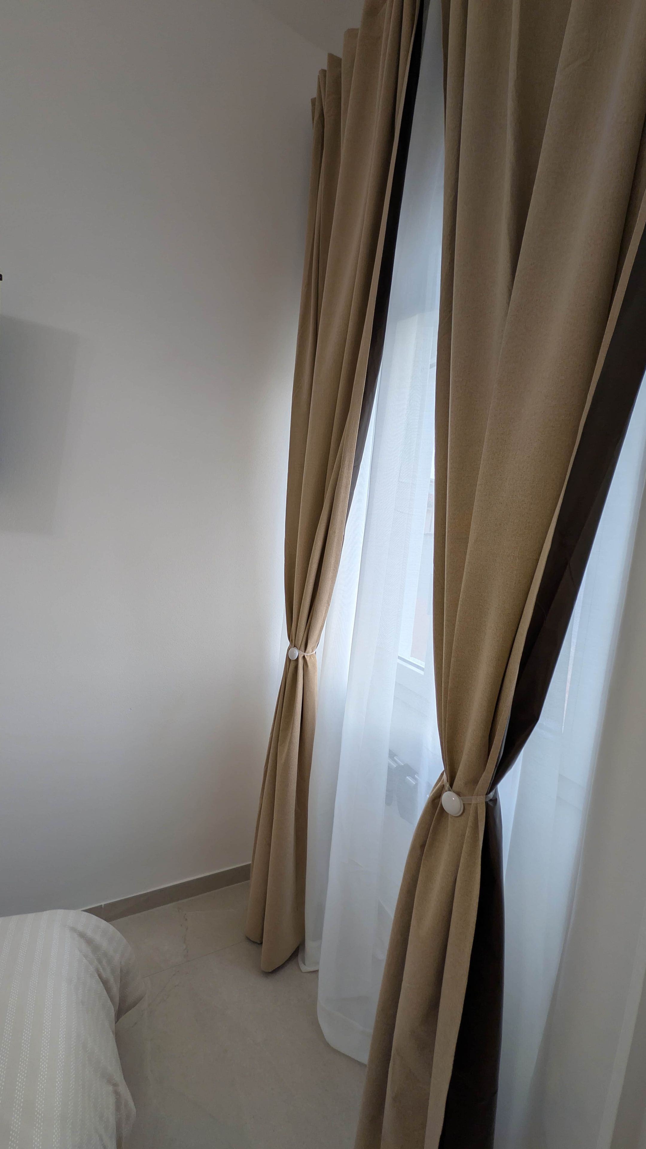 Una stanza con tende beige legate in basso, un letto bianco e una parete spoglia.