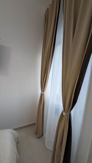 Una habitación con cortinas color beige atadas en la parte inferior, una cama blanca y una pared lisa.