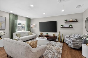 A modern living room with a sofa, armchair, and coffee table, a flat-screen TV mounted on the wall, and decorative items on shelves. - 3-bedroom house in beautiful St. Cloud with cool AC breeze (St. Cloud)