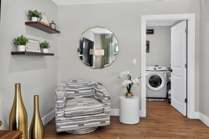 Ein Wohnzimmer mit einer Couch, einem runden Spiegel und einem kleinen Tisch mit einer Vase. Eine Tür führt zu einem Waschraum.