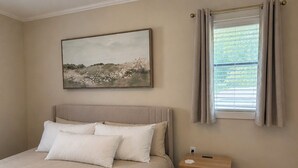 Een slaapkamer met een bed, nachtkastje, schilderij en een raam met gordijnen.