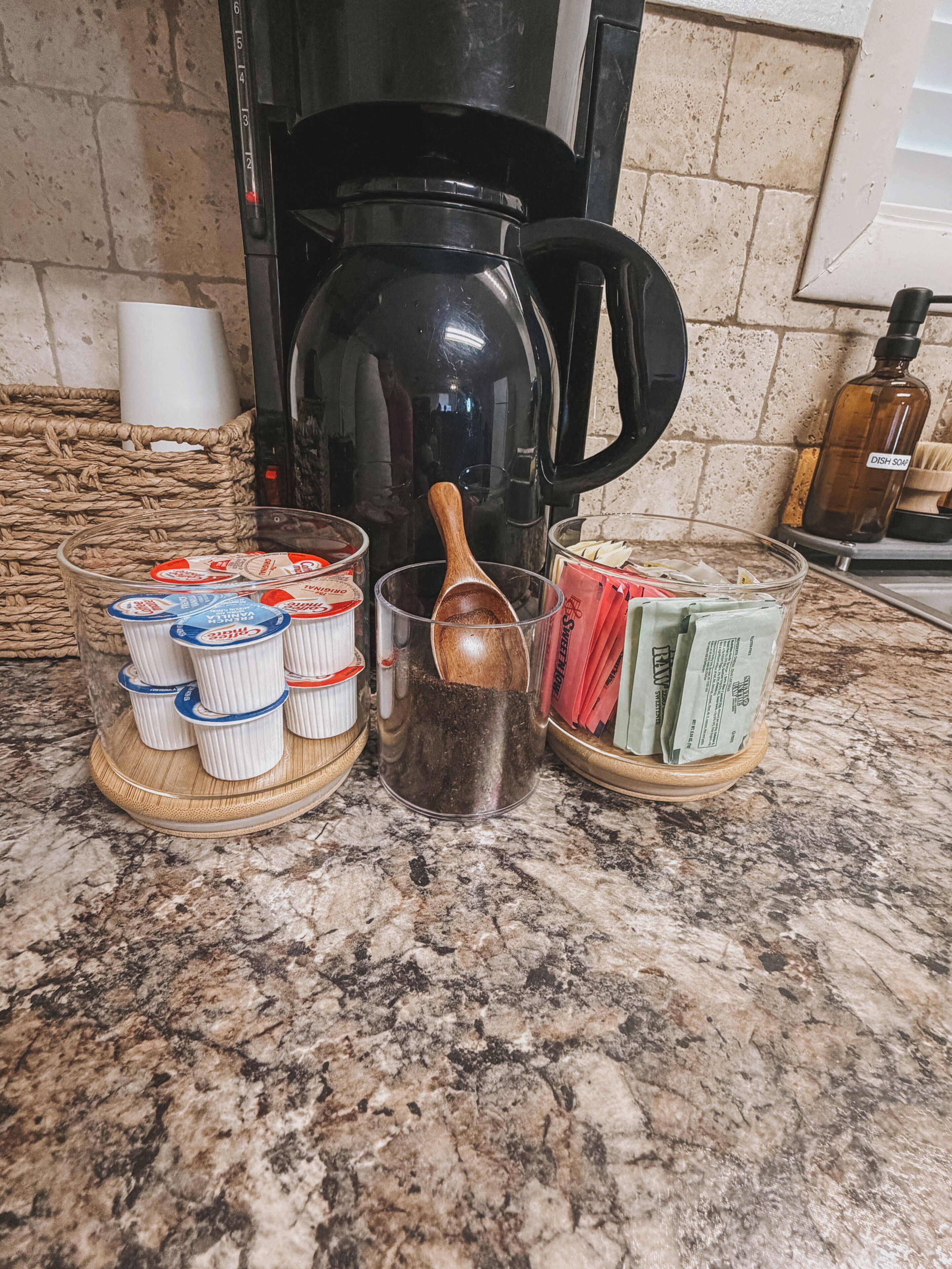 Un plan de travail de cuisine avec une machine à café, une cuillère en bois, un récipient en verre et un panier de serviettes en papier.