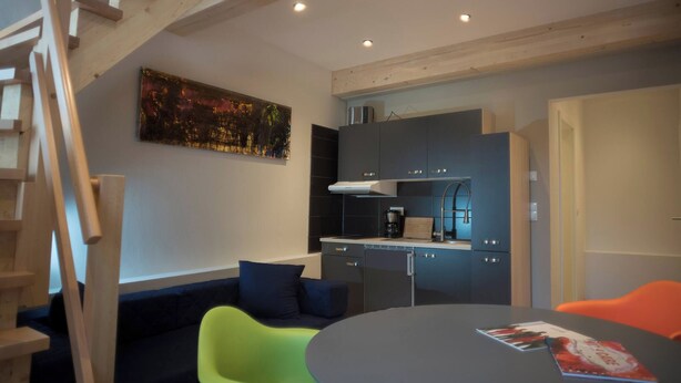 A modern kitchen with dark cabinets, a built-in oven, and a coffee machine. There is a dining table with a magazine on it, surrounded by colorful chairs. A staircase is visible in the background. - Maisonette Apartment The Haystack Ad1716 (Marktheidenfeld)