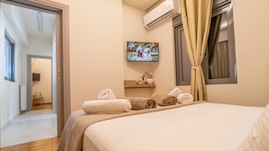 Quarto de hotel com cama, televisão fixada na parede, janela com cortinas e porta que dá para outro quarto.