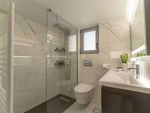 A modern bathroom with a glass-enclosed shower, a toilet, and a sink area with a mirror and a potted plant. - Nuvion (Athens)