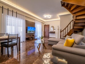 Una sala moderna con una televisión de pantalla plana, un comedor con mesa y sillas, un sofá con cojines, y una escalera.
