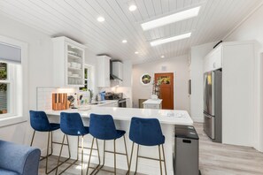Une cuisine moderne avec des meubles blancs, un îlot central doté de tabourets de bar, un réfrigérateur en acier inoxydable et une fenêtre avec des stores.