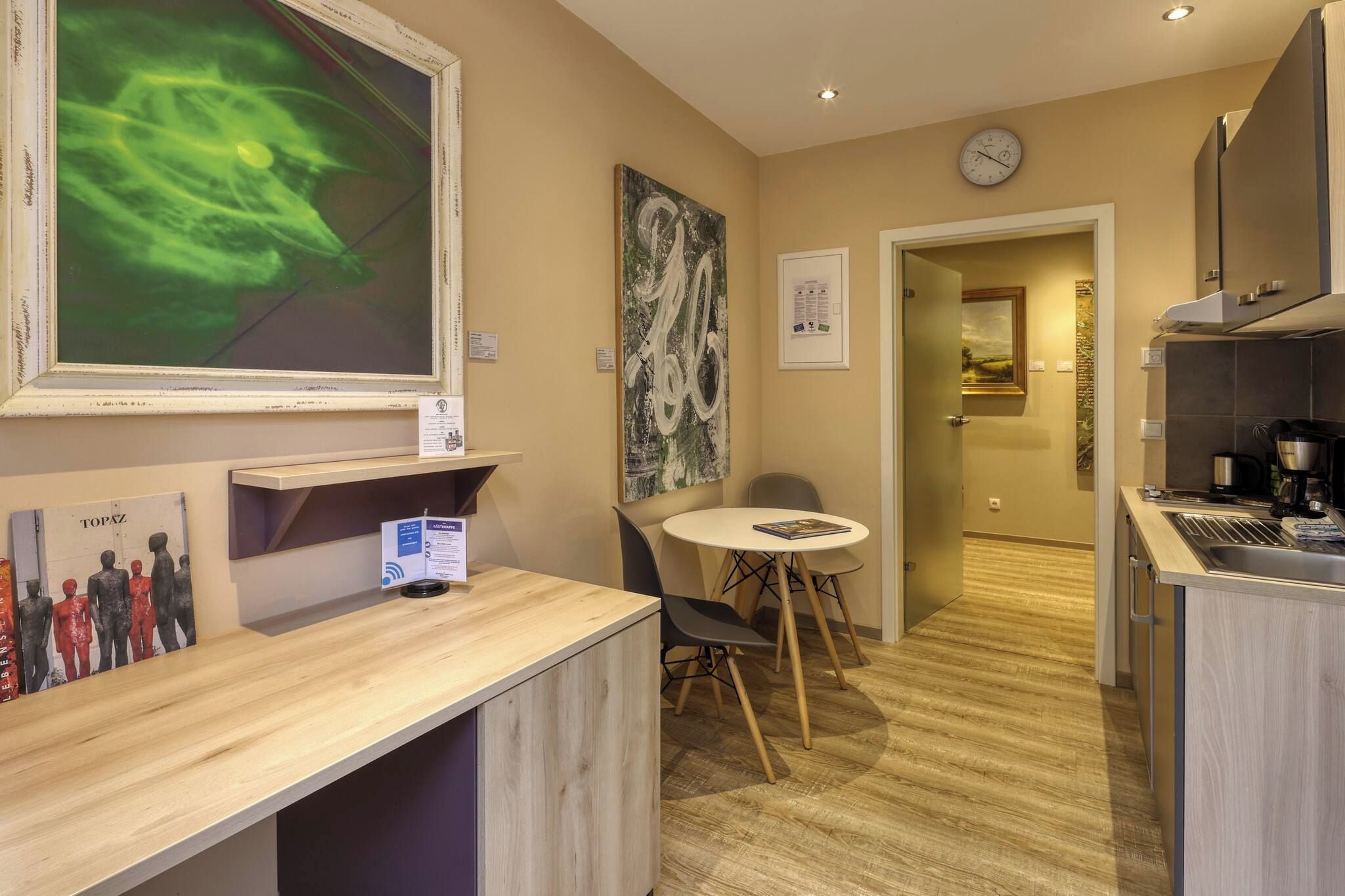 A hotel room with a wooden desk, a round table, a kitchen area, and artwork on the walls.