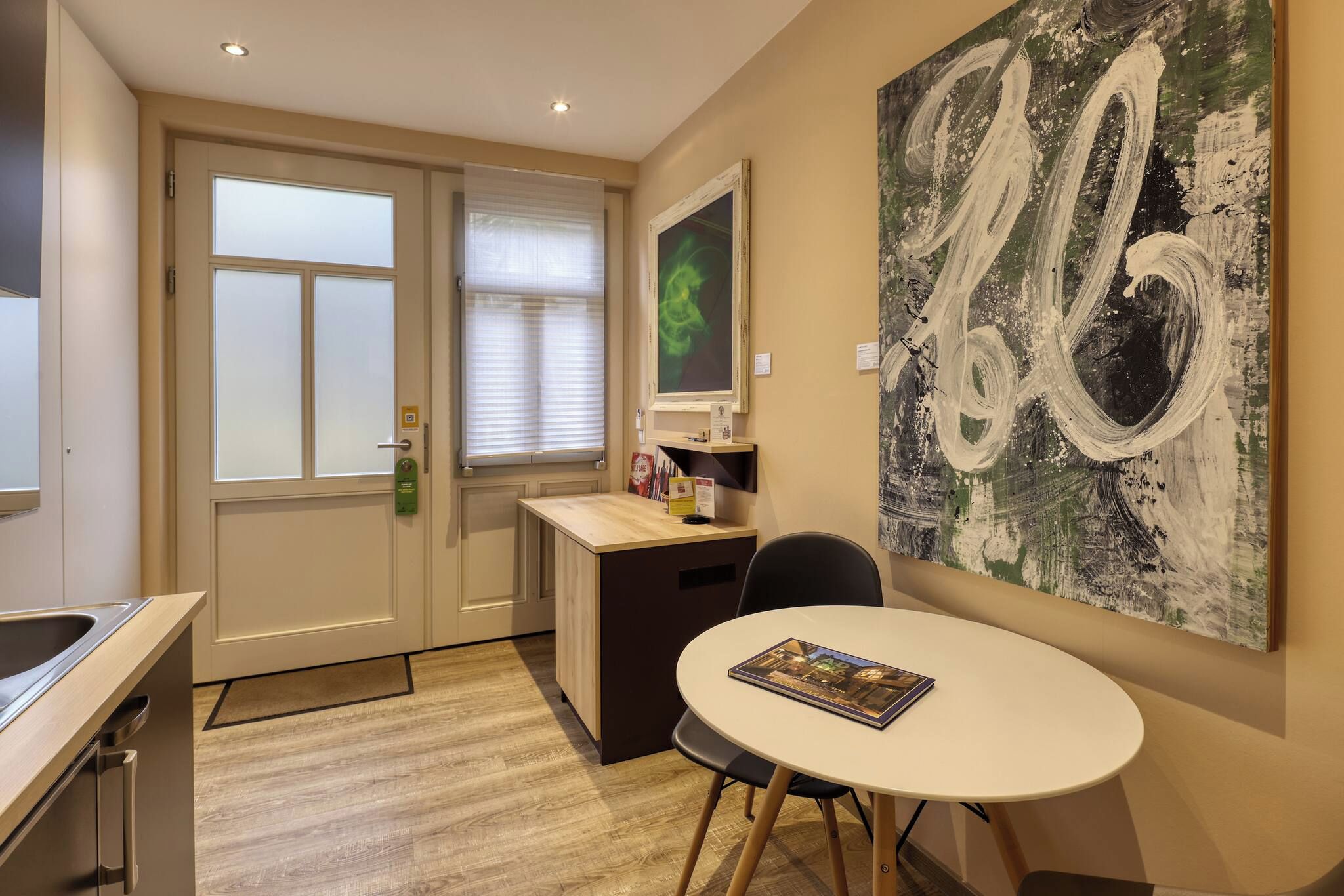 A hotel room with a desk, a chair, a dining table, a large abstract painting, and a window with blinds.