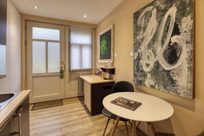 Quarto de hotel com escrivaninha, cadeira, mesa de jantar, uma pintura abstrata grande e janela com persianas.
