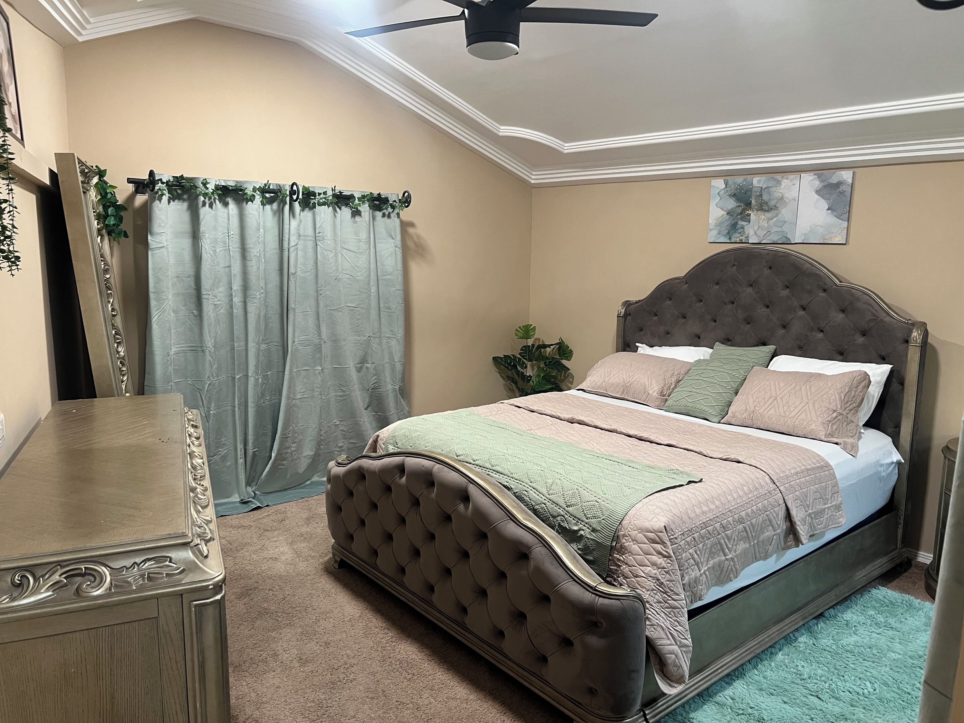A bedroom with a tufted headboard bed, a nightstand, a dresser, a ceiling fan, and a window with curtains.