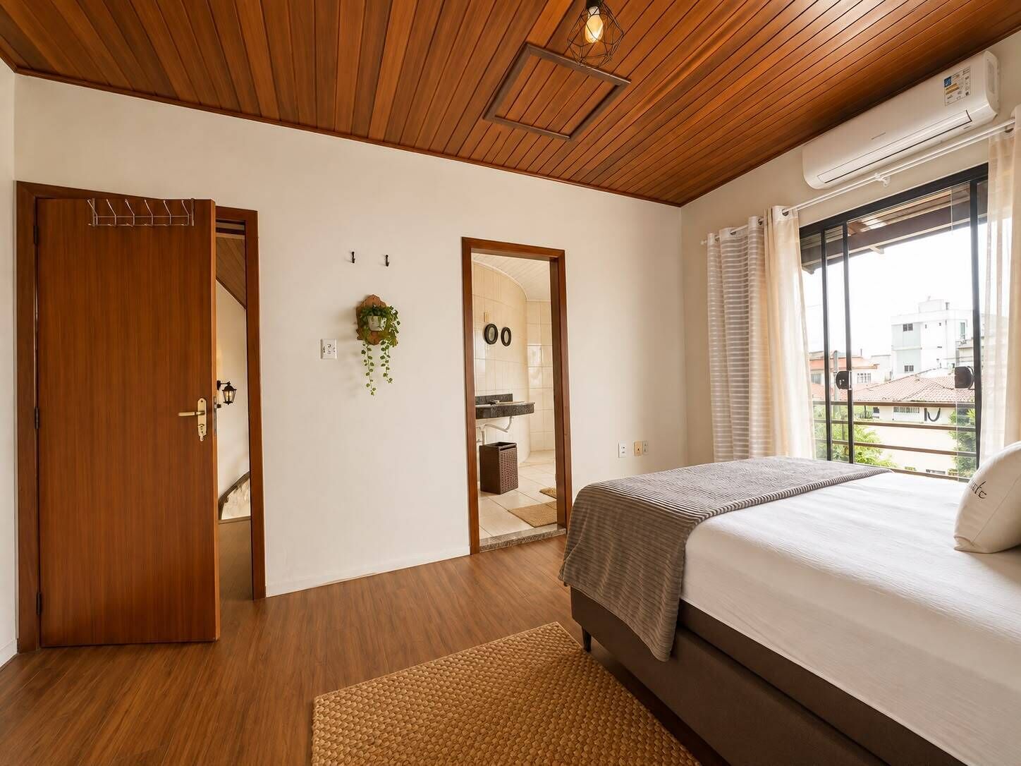 Quarto com cama, piso de madeira, porta, planta presa na parede e janela com vista.