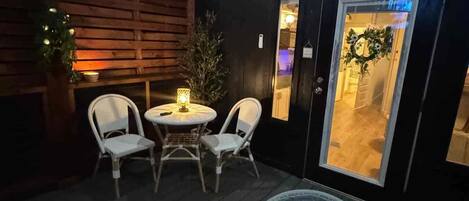 A wooden deck with a round table and two chairs, a door with a frosted glass panel, and a small plant.