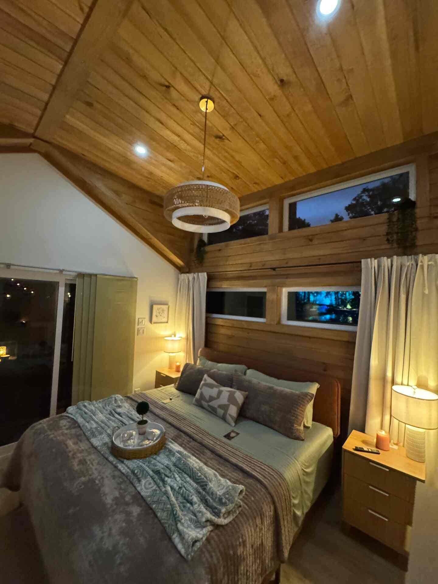 A bedroom with a sloped ceiling, a bed with a patterned bedspread, a bedside table with a lamp, and a window with curtains.
