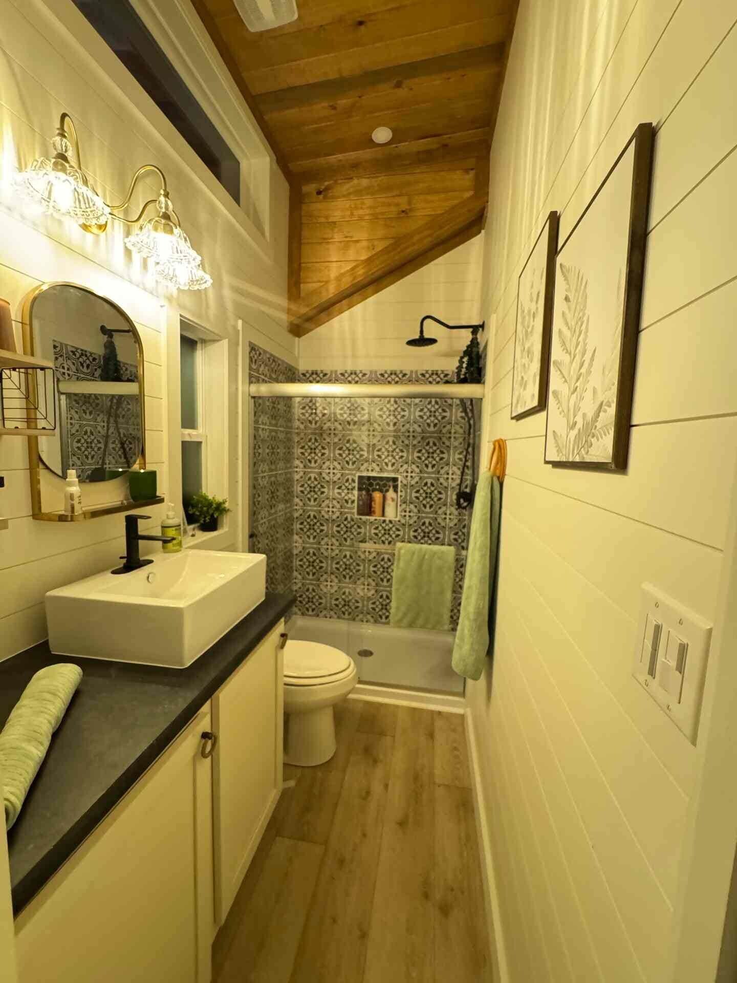 A narrow bathroom with a wooden ceiling, a white sink, a toilet, and a shower area with patterned tiles.