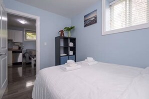 A bedroom with a bed, a small desk, a chair, a wardrobe, and a view of the kitchen and living area. - 2-Bedroom Suite. Walk to SkyTrain, Shops & Cafés (Vancouver)