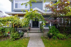 A two-story house with a front porch, a black door, and a stone foundation. - 2-Bedroom Suite. Walk to SkyTrain, Shops & Cafés (Vancouver)
