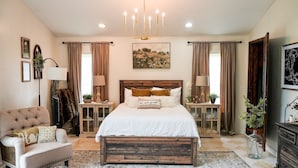 A bedroom with a large bed, two bedside tables, a nightstand, a chair, a wooden headboard, a chandelier, and a painting on the wall. - Cozy 4-bedroom cabin in tranquil Newcomerstown with AC (Newcomerstown)