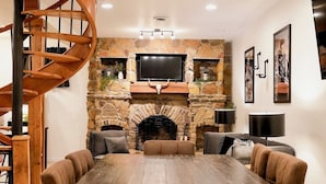 A spacious room with a large wooden dining table surrounded by chairs, a stone fireplace, a mounted flat-screen TV, and a spiral staircase. - Cozy 4-bedroom cabin in tranquil Newcomerstown with AC (Newcomerstown)