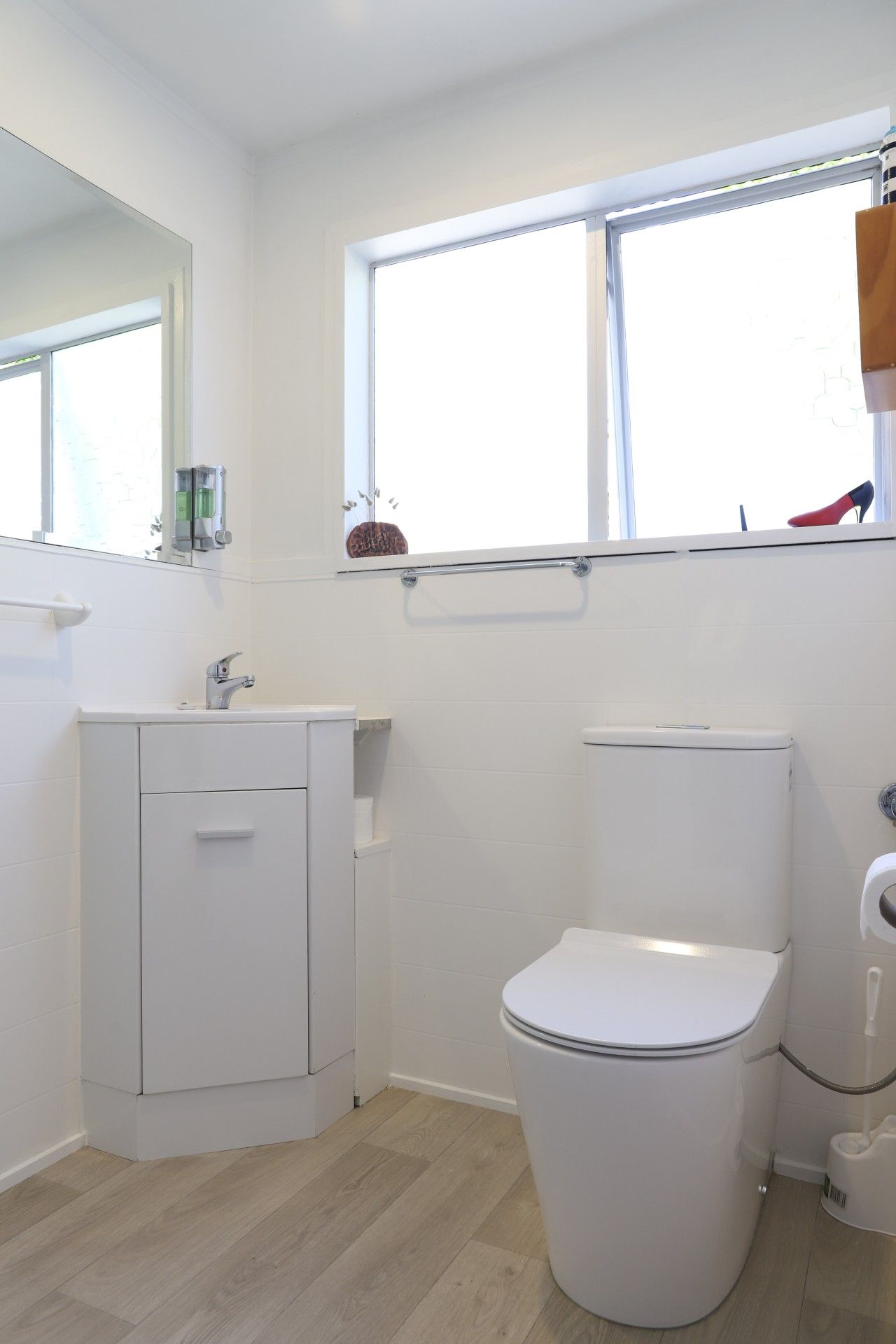 A small, white bathroom with a toilet, a sink, and a mirror. There is a window above the toilet, and a shelf with a decorative item.