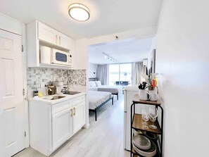 A compact hotel room with a kitchenette, a bed, a dining area, and a view of the city. - Cozy Studio w/ Pool View  2 Queens & Patio Dining (Honolulu)