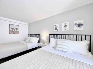 A hotel room with a large bed, a smaller bed, a nightstand with a lamp, and three wall-mounted artworks. - Cozy Studio w/ Pool View  2 Queens & Patio Dining (Honolulu)