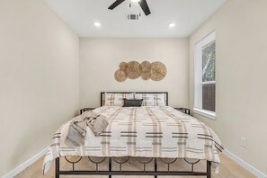 Ein Schlafzimmer mit einem Bett, einem Deckenventilator, Einbauleuchten, einem Wandlüfter und einem Fenster mit Jalousien.