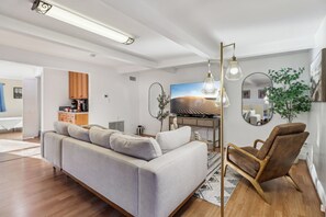 A modern living room with a sofa, a wooden chair, a TV, and a mirror on the wall. - Entire Private Home w/ 4 Beds, 2 Bath, Gated Yard, 2 Living Rooms, Ranch Style (Spring Valley)