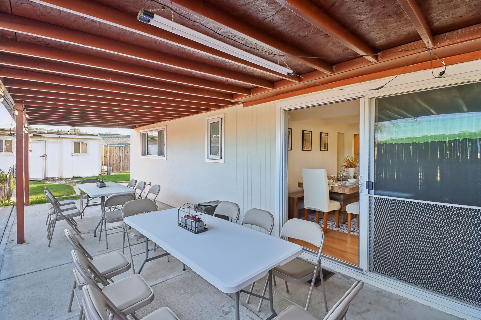 Un patio al aire libre con mesa y sillas, un área cubierta con vigas de madera y una puerta corrediza que da a un espacio interior.