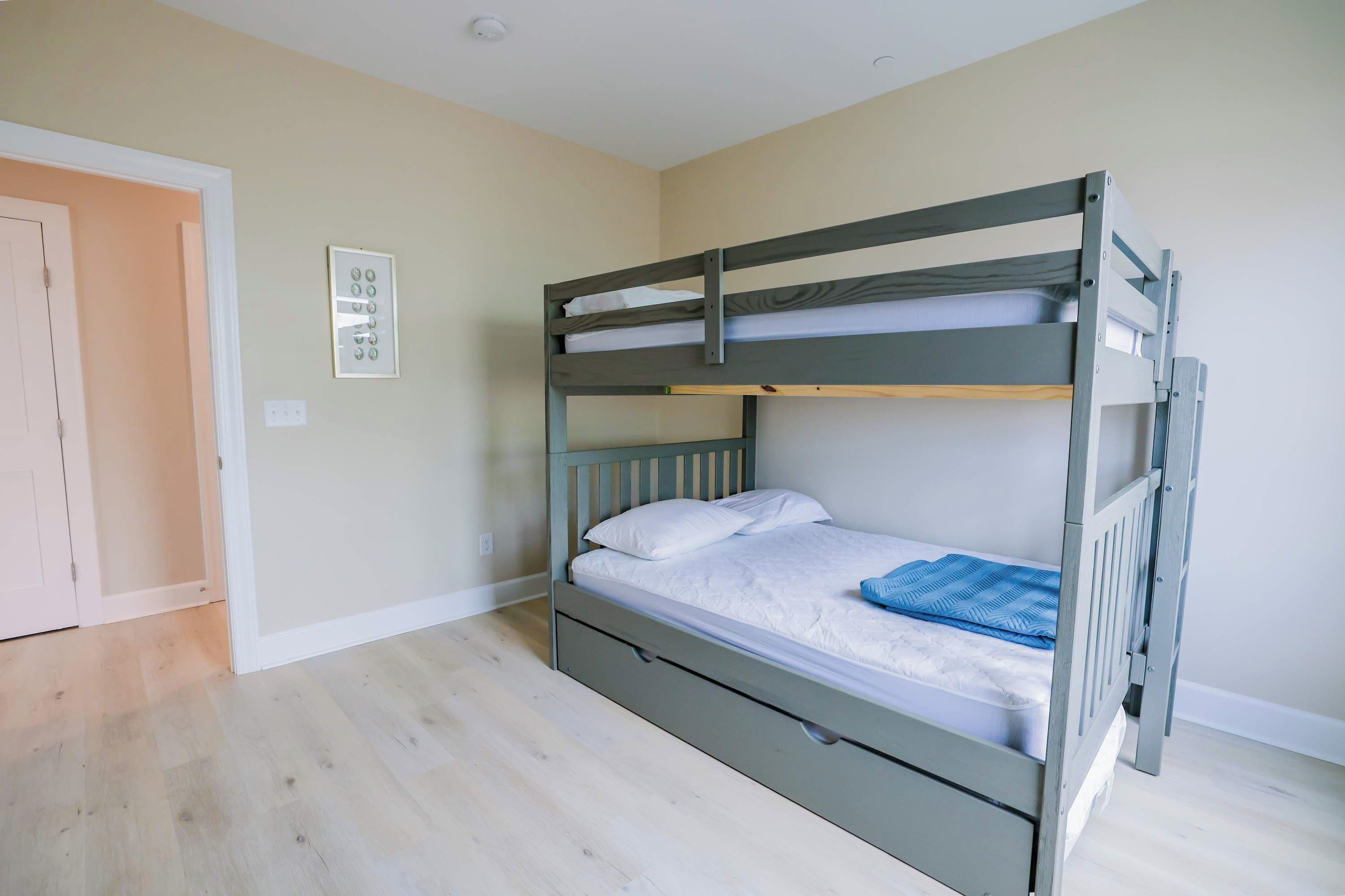 Un lit superposé avec un lit inférieur, un matelas blanc et une couverture bleue. Il y a une porte à gauche et un interrupteur sur le mur.