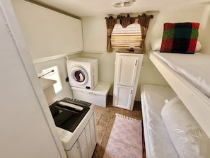 A compact room with a washing machine, a small cabinet, a bed, and a window with curtains.