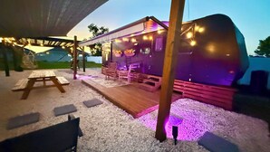 Un espace extérieur aménagé avec des tables et des chaises, protégé par un auvent, ainsi qu’une terrasse en bois et des guirlandes lumineuses.