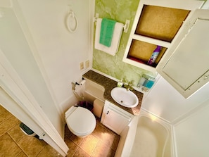 Ein Badezimmer mit Toilette, Waschbecken und Badewanne. Ein Handtuch hängt an der Wand und ein kleines Regal mit Toilettenartikeln ist vorhanden.