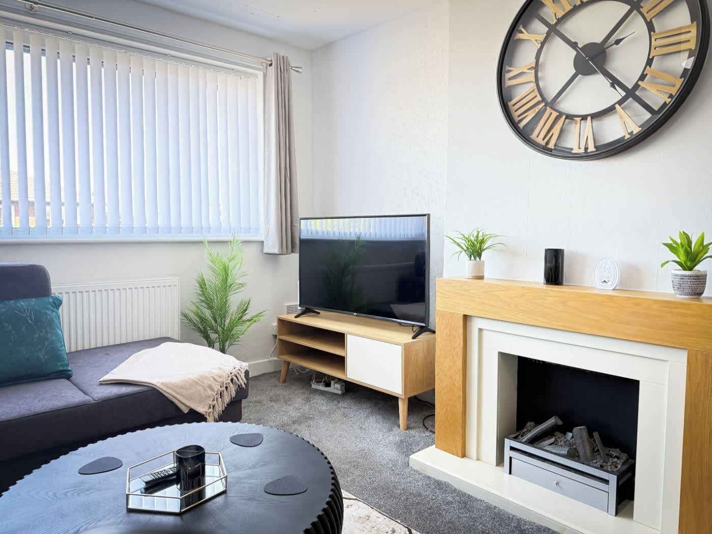 A modern living room with a large wall clock, a flat-screen TV, a sofa, a coffee table, and a fireplace.