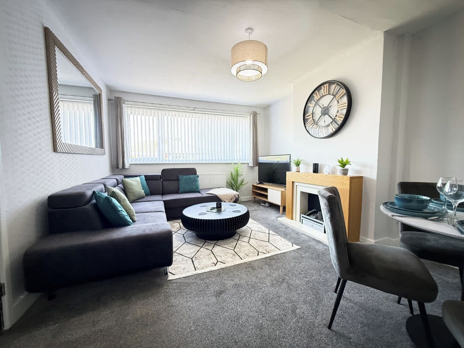 Een moderne woonkamer met een hoekbank, een rond salontafeltje, een open haard, een eethoek met een tafel gedekt voor twee personen, en een grote wandklok.