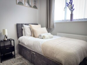 A bedroom with a bed, bedside table, and a window with curtains. - Cosy Southport Bungalow | 2 Bedroom with Parking (Banks)