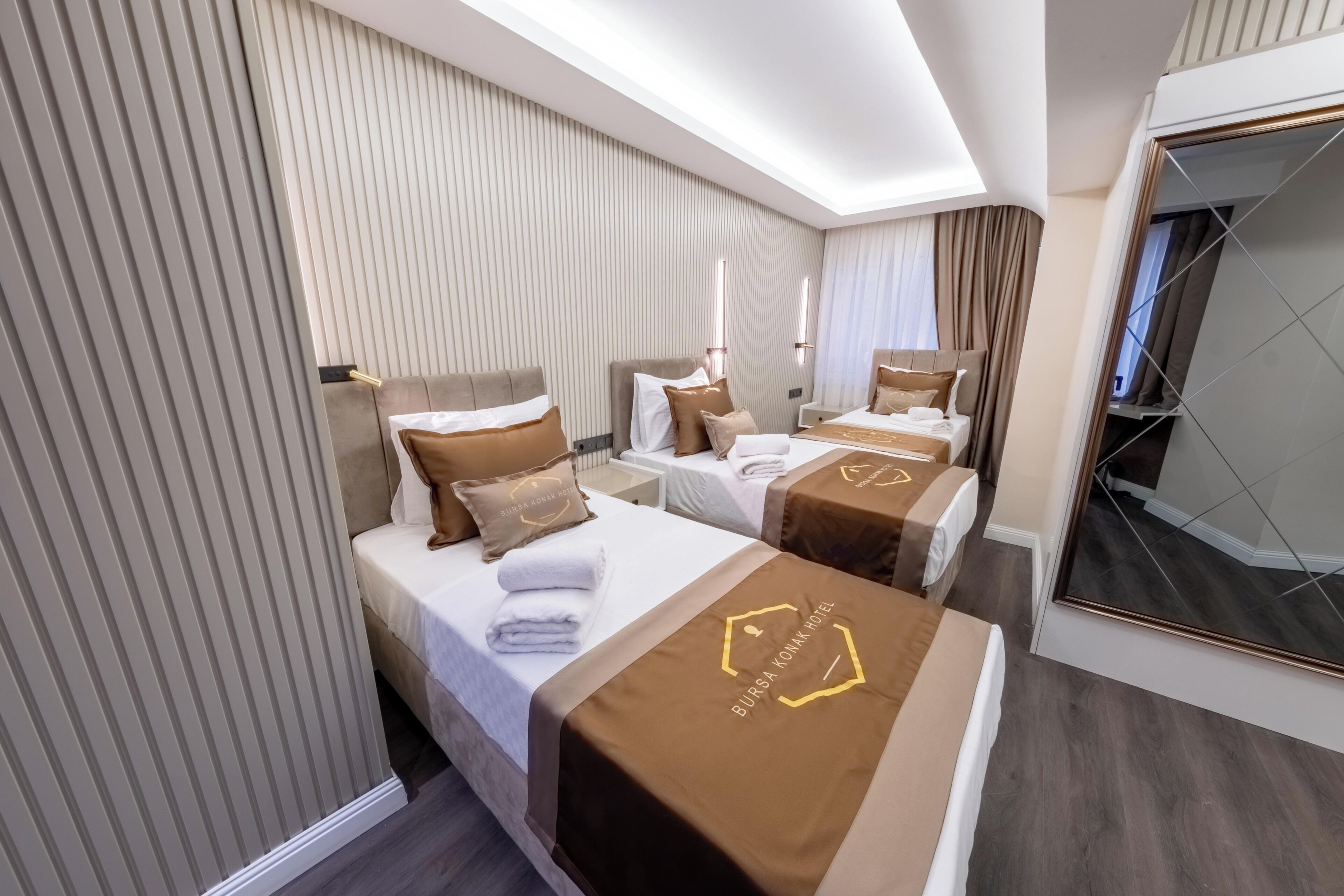 Et hotelværelse med tre senge, hver med et brunt sengegavl og et hvidt dynebetræk. Værelset har et moderne design med et stort spejl og udsigt over byen.