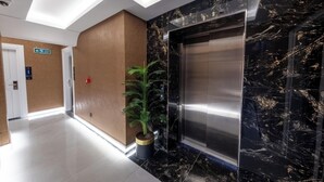 Ascensor moderno con detalles de mármol, una planta en maceta y señalización de salida clara en un pasillo con suelo claro.
