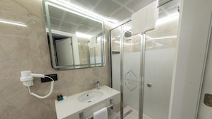 Un baño moderno con un lavamanos blanco, un secador de pelo, cabina de ducha con mampara de vidrio y un espejo con luz en el techo.