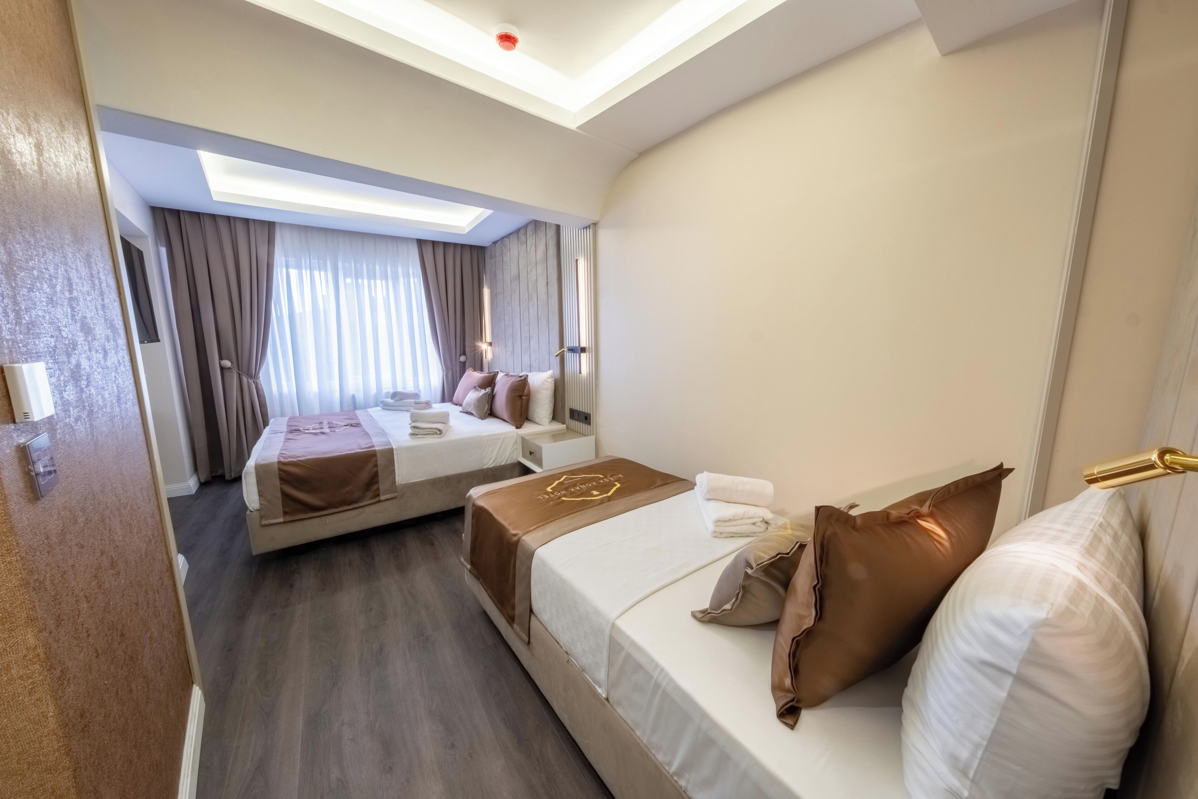 Habitación de hotel con dos camas, un ventanal con cortinas, una lámpara en el techo y un televisor montado en la pared.