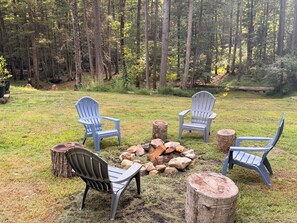 An outdoor seating area with Adirondack chairs around a fire pit, surrounded by a grassy field and a forested backdrop.