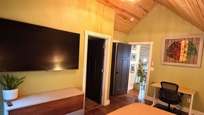 Une chambre à coucher comprenant un lit, une commode, un téléviseur et un bureau avec une chaise. Une porte donne accès à une autre pièce.