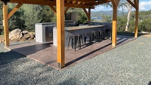 Une cuisine extérieure avec un coin bar aménagé sous une pergola en bois, entourée de gravier et d’arbres.