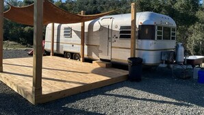 A recreational vehicle (RV) parked on a gravel surface with a wooden platform and a canopy structure. - Moonshadow Flat Glamping: Gypsum Tent (Penn Valley)